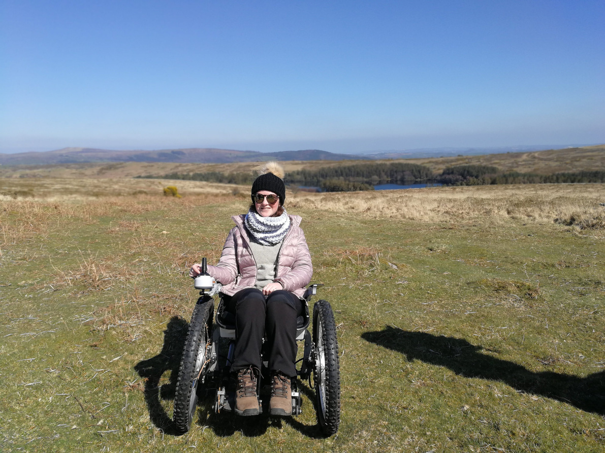 Image of Bailey in their all terrain wheelchair at Dartmoor. There is blue skies and rolling hills behind Bailey. 